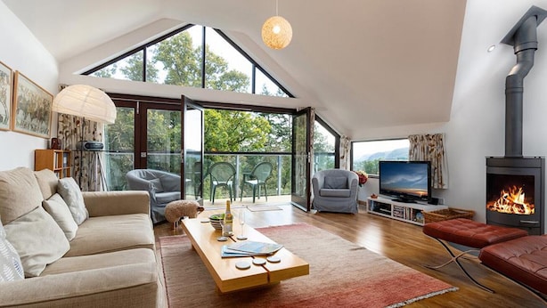 Ein geräumiges Wohnzimmer mit hoher Decke, Kamin, Fernseher und großem Fenster mit Blick auf Grünflächen.
