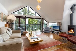 A spacious living room with a high ceiling, a fireplace, a television, and a large window overlooking greenery. - Hafod Rowen - Four Bedroom Cottage, Sleeps 8 (Rowen)