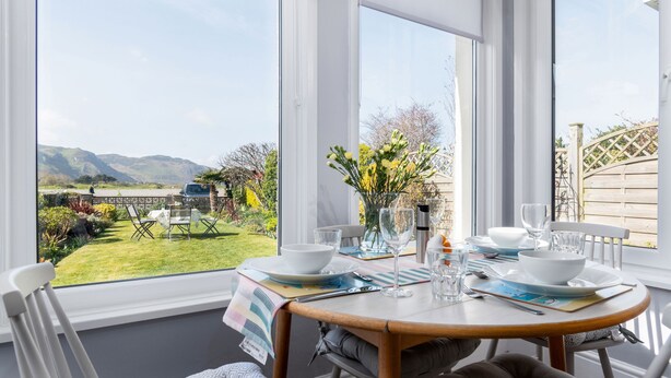 A dining area with a table set for two, a vase with flowers, and a view of a garden and hills. - Sandbanks - Two Bedroom Cottage, Sleeps 3 (Deganwy)