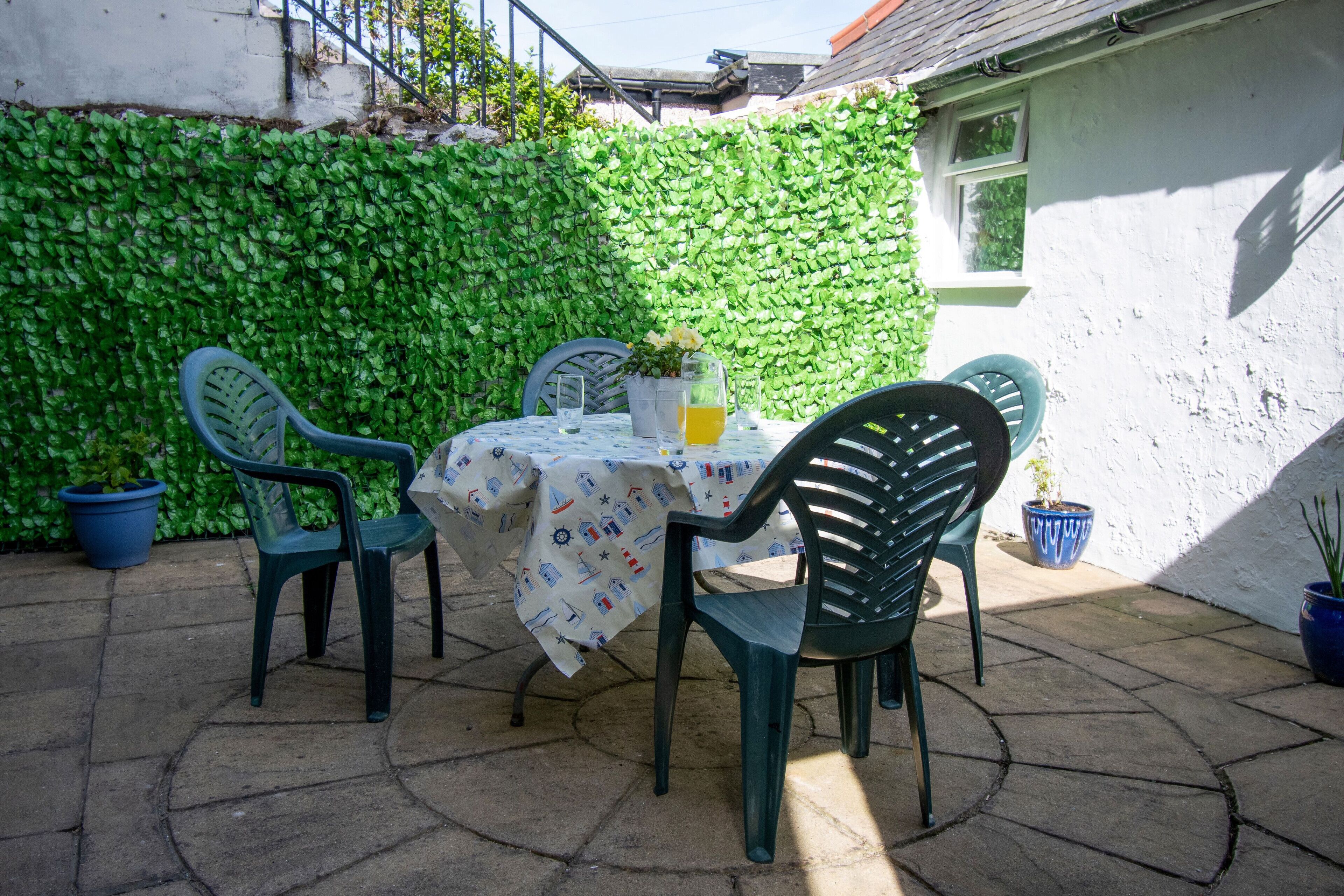 Eine Terrassengruppe im Freien mit Tisch und Stühlen, umgeben von einer grünen, blätterreichen Wand und Topfpflanzen.