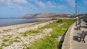 A coastal promenade with benches, a pebble beach, and a distant mountain range. - Sandbanks - Two Bedroom Cottage, Sleeps 3 (Deganwy)