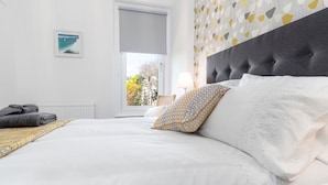 A neatly made bed with white linens and patterned pillows, a gray headboard, and a view of the outdoors through a window. - Sandbanks - Two Bedroom Cottage, Sleeps 3 (Deganwy)