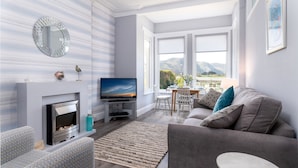 A modern living room with a fireplace, a television, a sofa, and a dining area with a view of mountains. - Sandbanks - Two Bedroom Cottage, Sleeps 3 (Deganwy)