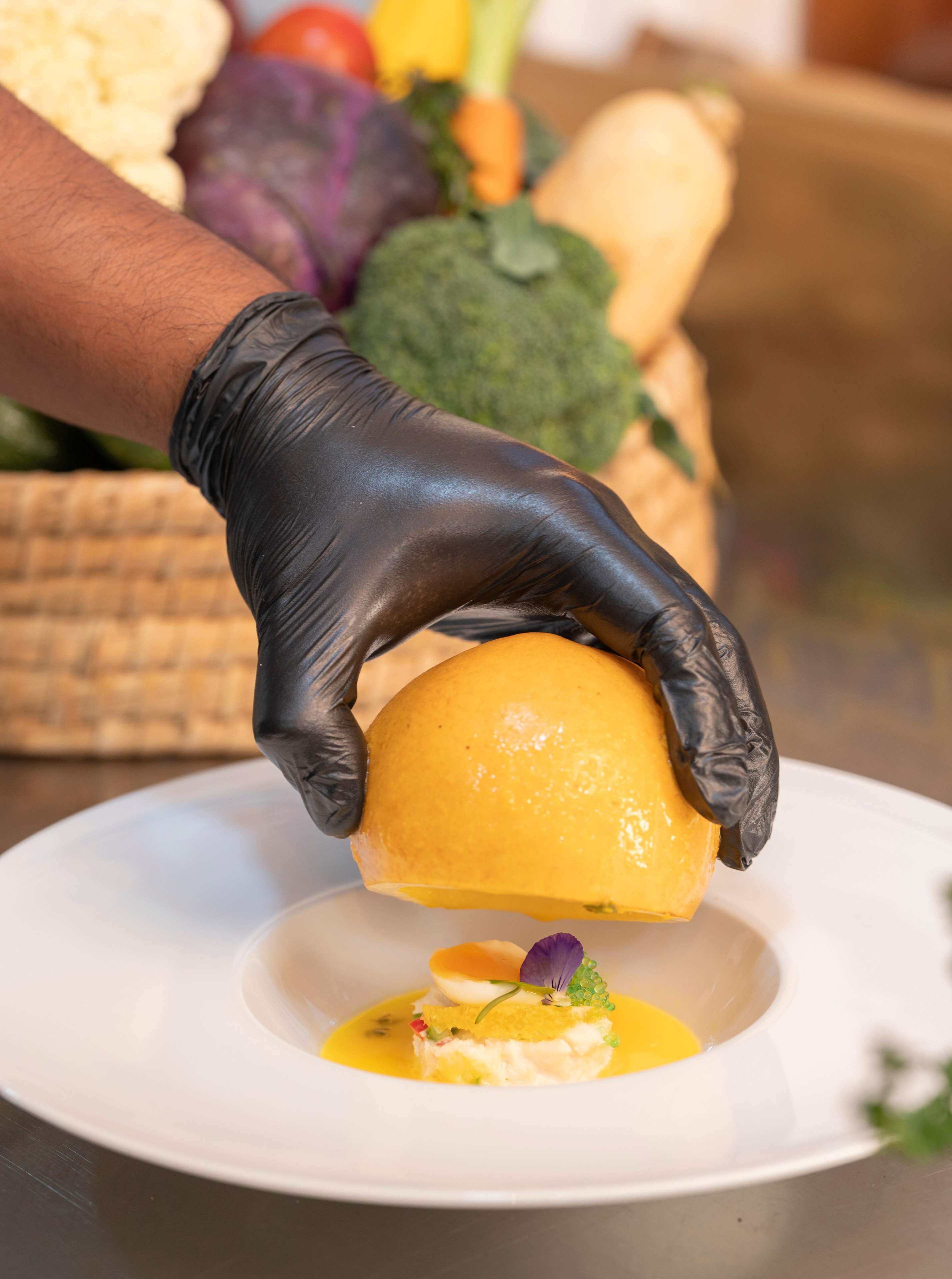 A chef wearing black gloves holding a lemon over a plate with a white sauce and a garnish.
