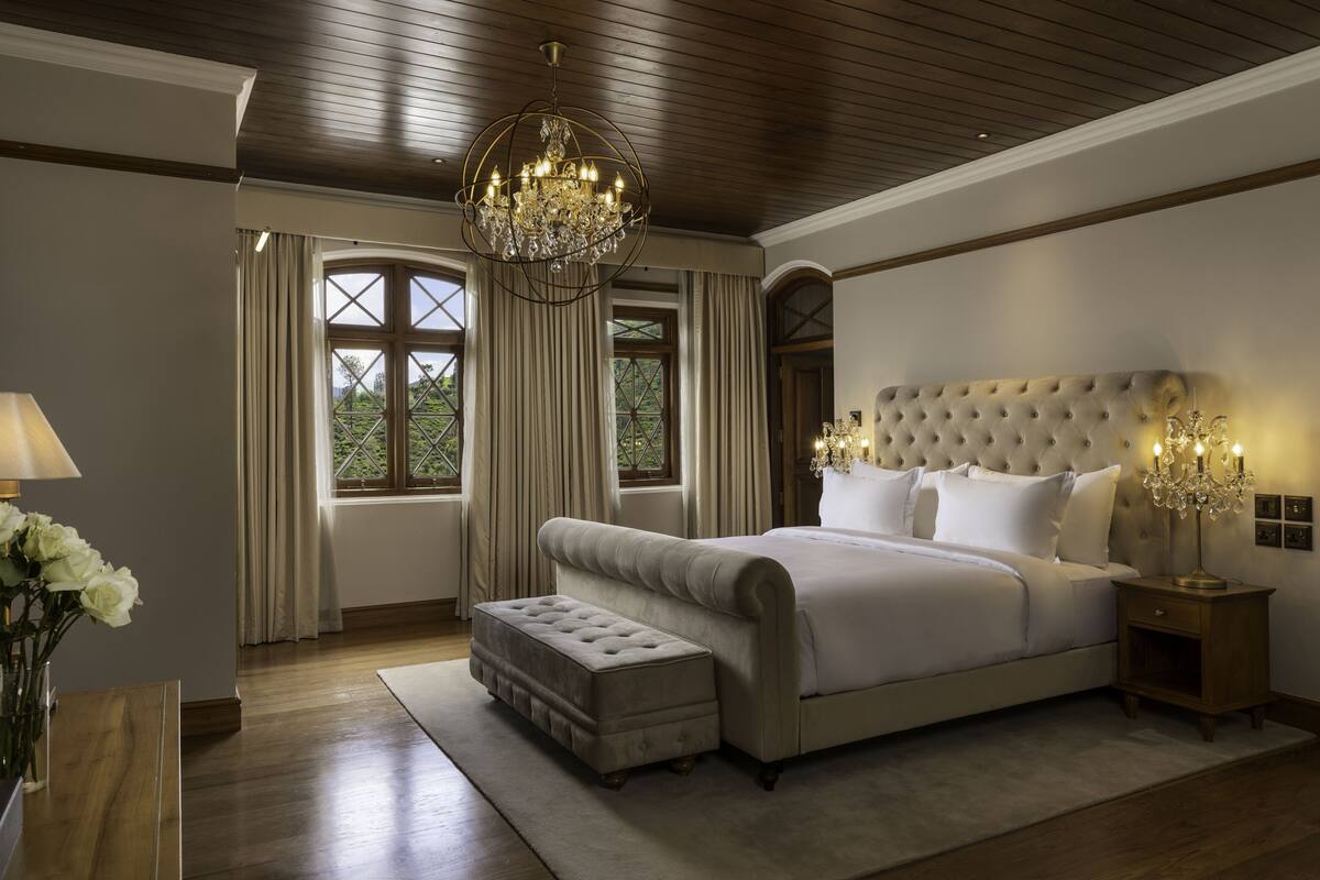 Ein Schlafzimmer mit einem großen Bett, einer Kristallleuchtre und Blick auf Grün hinter den Fenstern.