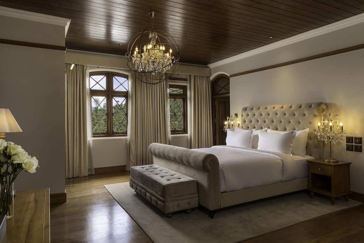 A bedroom with a large bed, a chandelier, and a view of greenery through the windows.