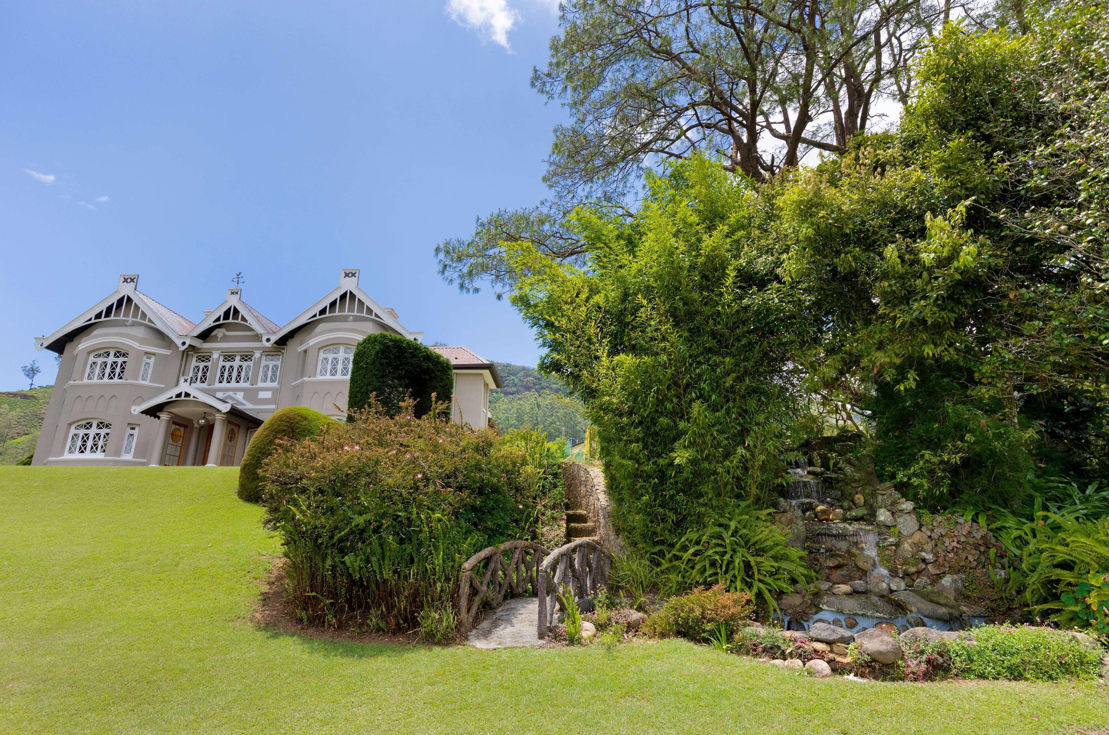 A large, two-story house with a prominent front entrance, surrounded by lush greenery and a small stone bridge.