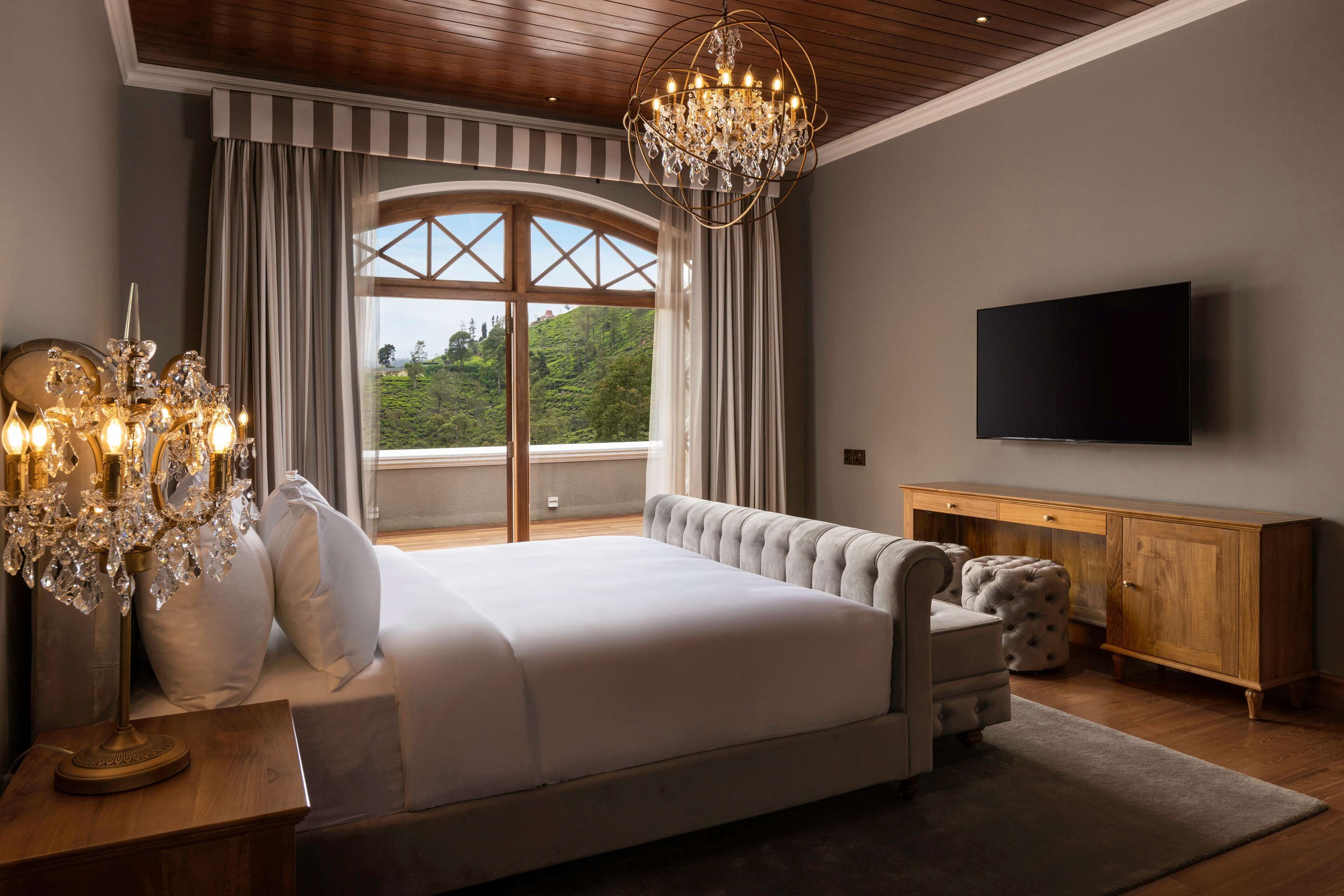 Ein Schlafzimmer mit einem großen Bett, einer Chandelier, einem Fernseher und Blick auf Grün durch ein Fenster.