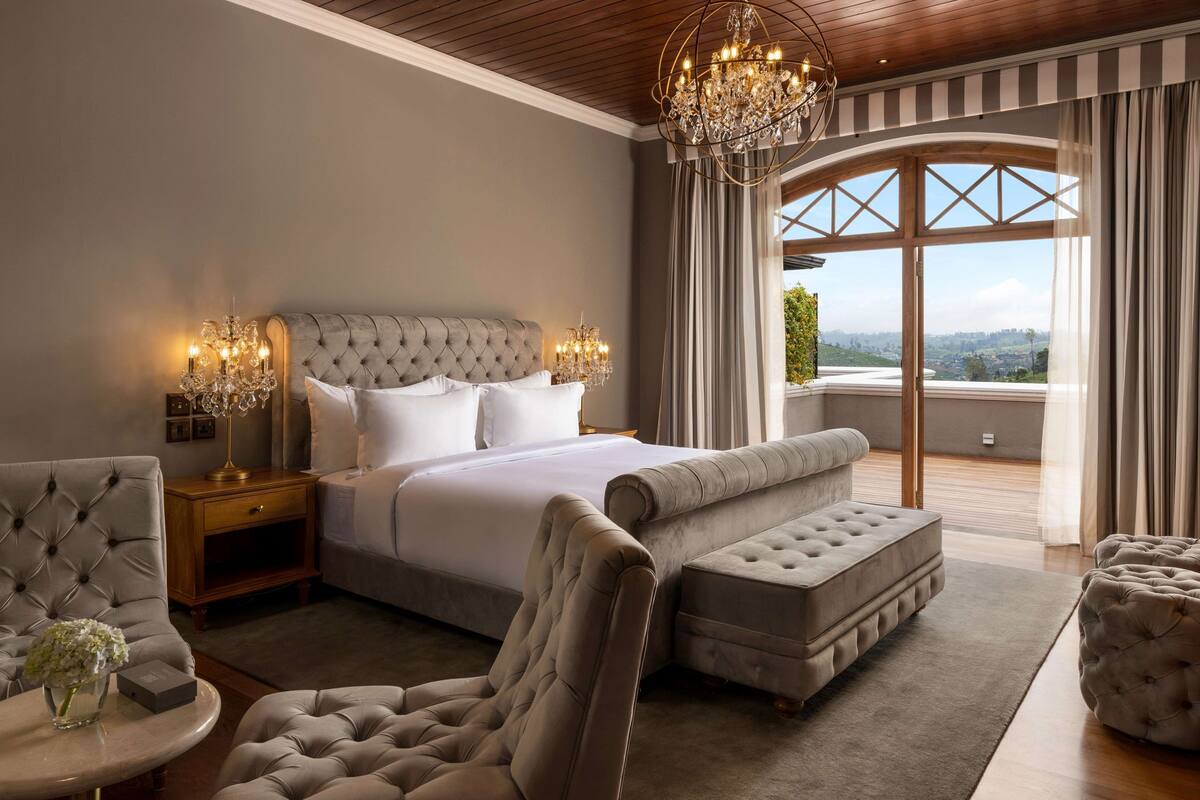 Ein Schlafzimmer mit einem großen Bett, einer Chandelier, einem Ausblick durch ein Fenster und einer Sitzecke mit Stuhl und Hocker.