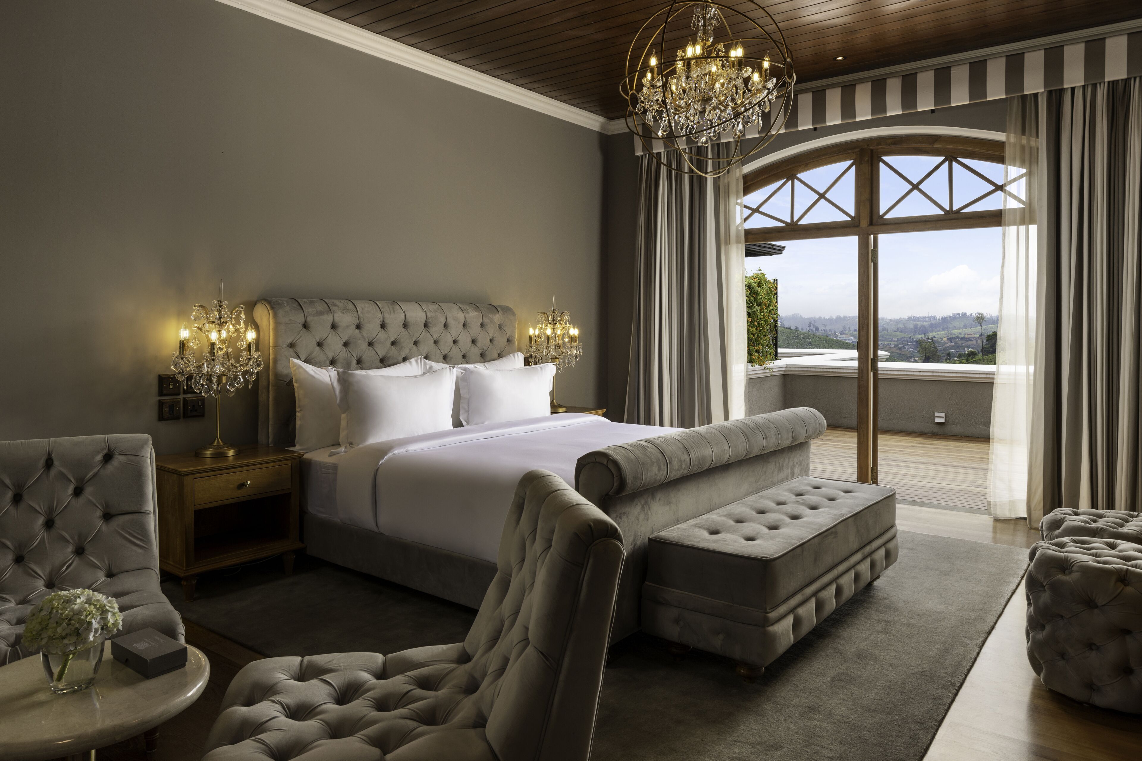 Une chambre à coucher avec un grand lit, un lustre, une vue par la fenêtre et un coin salon comprenant un fauteuil et un pouf.
