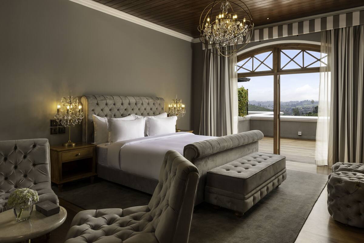 Ein Schlafzimmer mit einem großen Bett, einer Chandelüster, einem Ausblick durch ein Fenster und einer Sitzecke mit Sessel und Hocker.