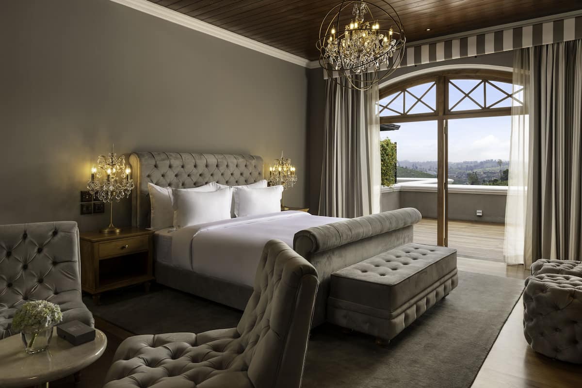 A bedroom with a large bed, a chandelier, a view through a window, and a seating area with a chair and ottoman.