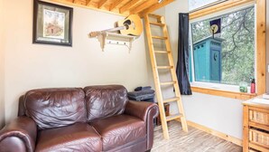 Eine Holzbalkendecke, ein braunes Ledersofa, eine Gitarre, eine Leiter, ein Fenster mit Blick auf Bäume und ein gerahmtes Bild an der Wand.