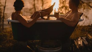 Zwei Personen in einer Badewanne im Freien, im Hintergrund ein Sonnenuntergang.