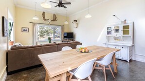 Ein Essbereich mit einem Holztisch, weißen Stühlen und einem braunen Sofa. Es gibt einen weißen Schrank mit Glastüren und einen Deckenventilator.
