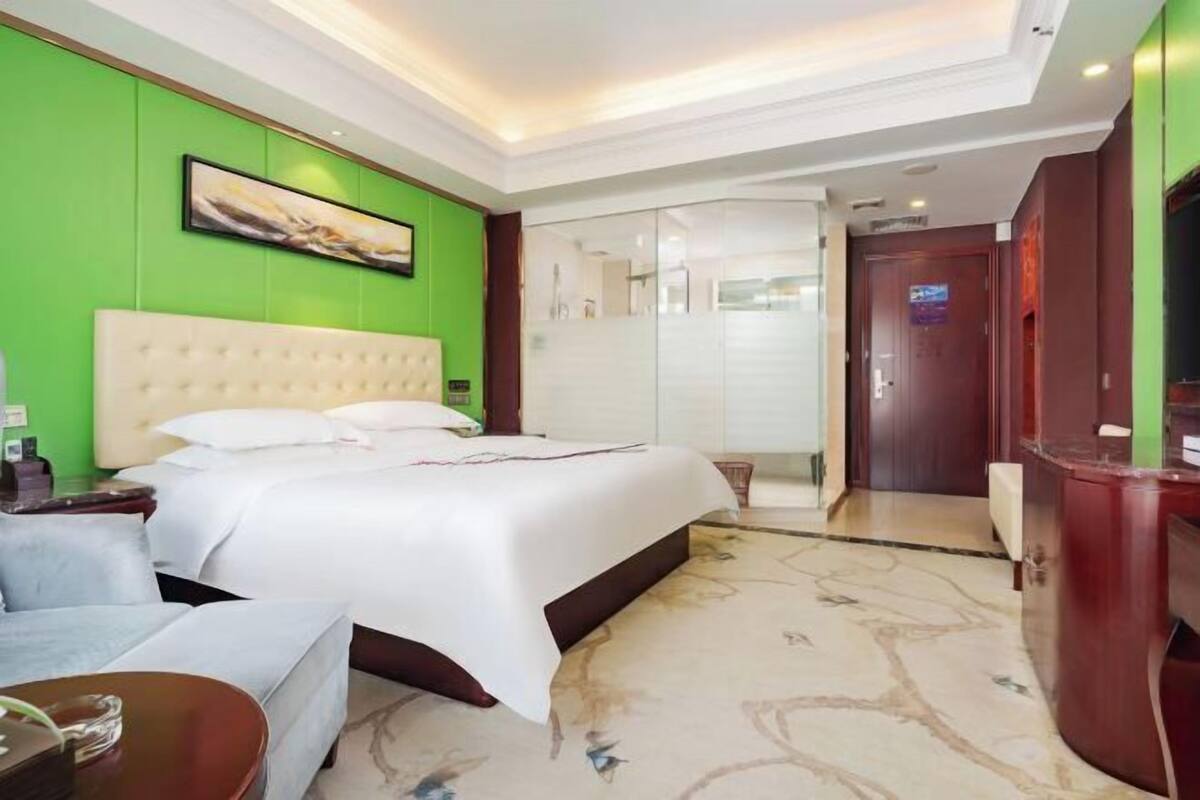 A hotel room with a large bed, a desk, a chair, a television, and a bathroom with a glass door.