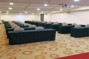 Un salón de conferencias con filas de mesas y sillas, piso alfombrado y una pantalla de proyector.