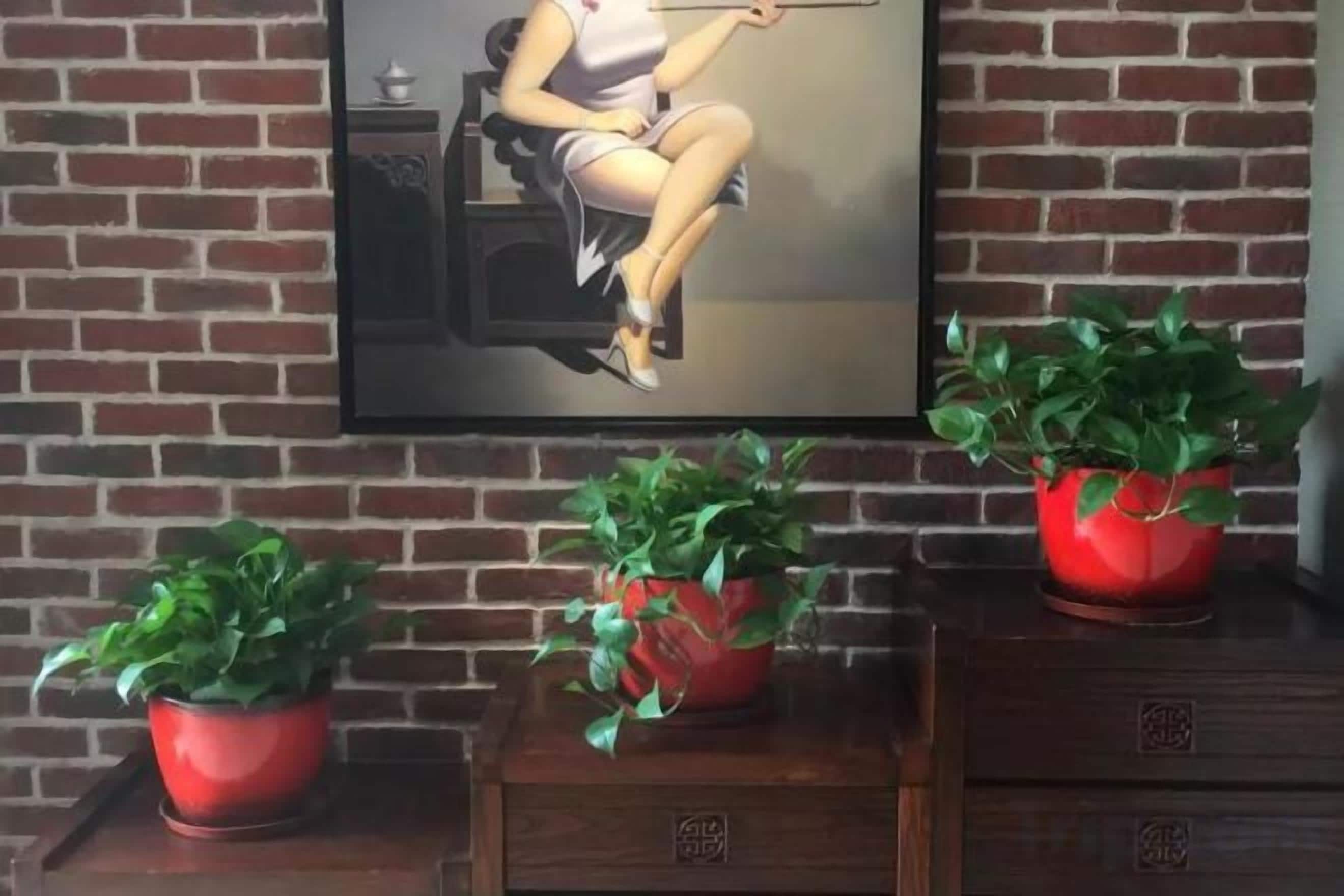 A brick wall with a framed painting of a woman, three red potted plants, and a wooden cabinet with drawers.