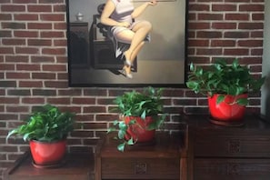 A brick wall with a framed painting of a woman, three red potted plants, and a wooden cabinet with drawers. - Huaying Wanrun Hotel (Cenxi)