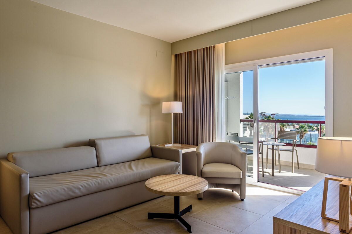 Una habitación de hotel moderna con un sofá, una mesa redonda pequeña, una silla y un balcón con vistas.