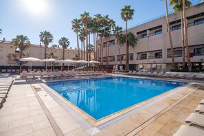 Una piscina con sillones de descanso y sombrillas, rodeada de palmeras y un edificio de varios pisos.