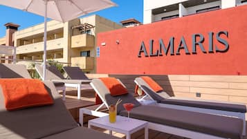 Un área de lounge en la azotea con sillones de sol, una mesa con una bebida y un edificio con la señalización 'ALMARIS'.