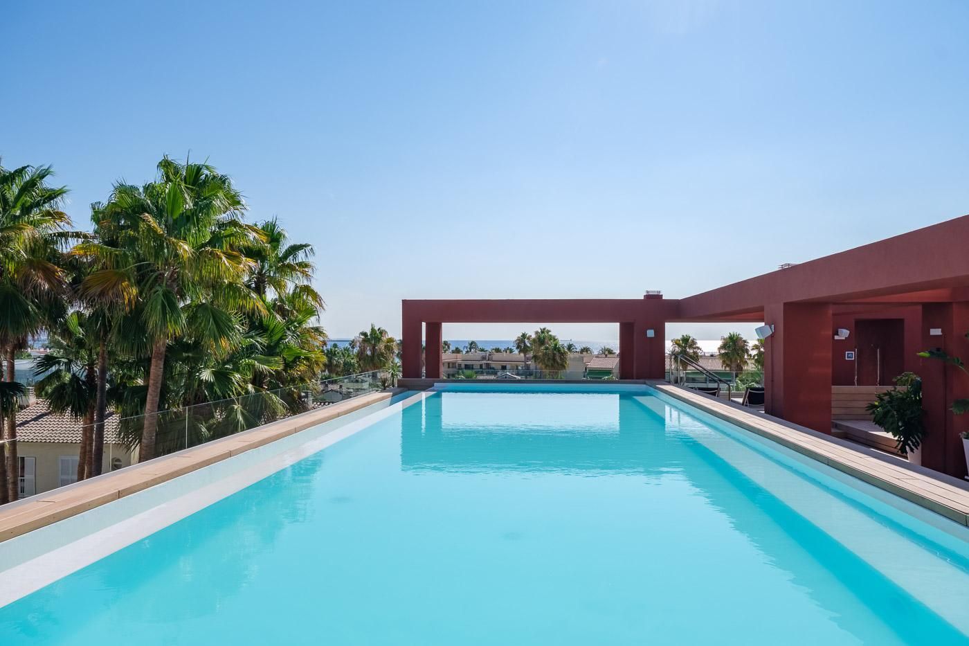 Una piscina azul cristalina con una estructura roja al fondo, rodeada de palmeras.