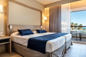 Habitación de hotel con dos camas, un balcón con mesa y sillas, y vistas a palmeras y al mar.