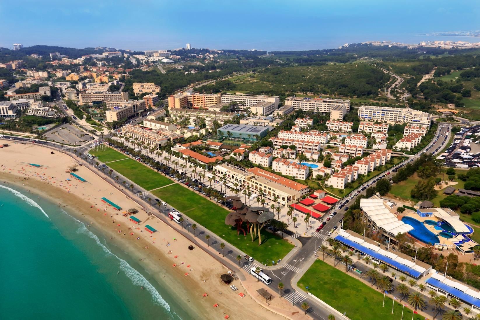 Vista aérea de un complejo turístico frente al mar, con varios edificios de hotel y una plaza central.