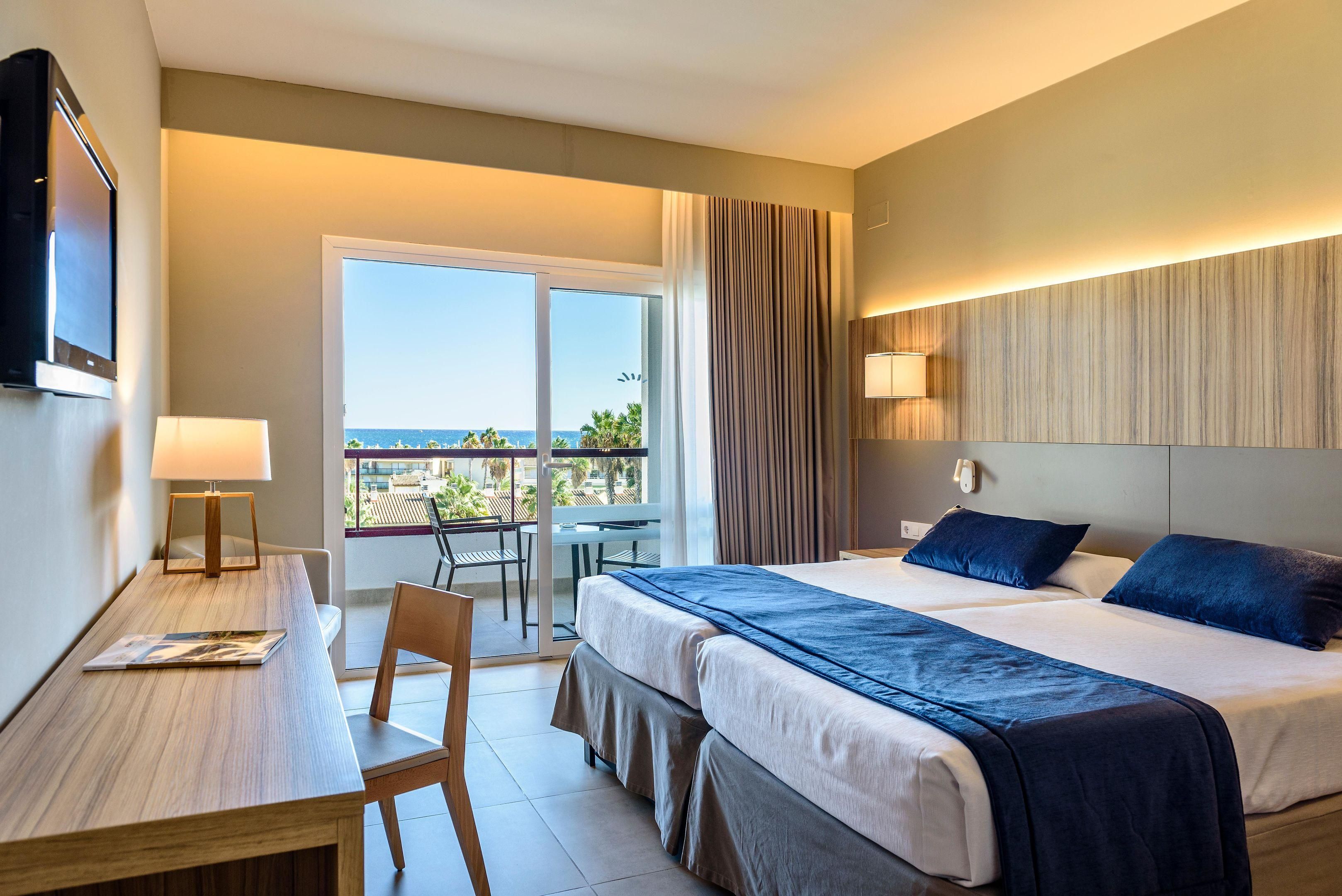 Habitación de hotel con cama, escritorio, silla, televisión y balcón con vistas.