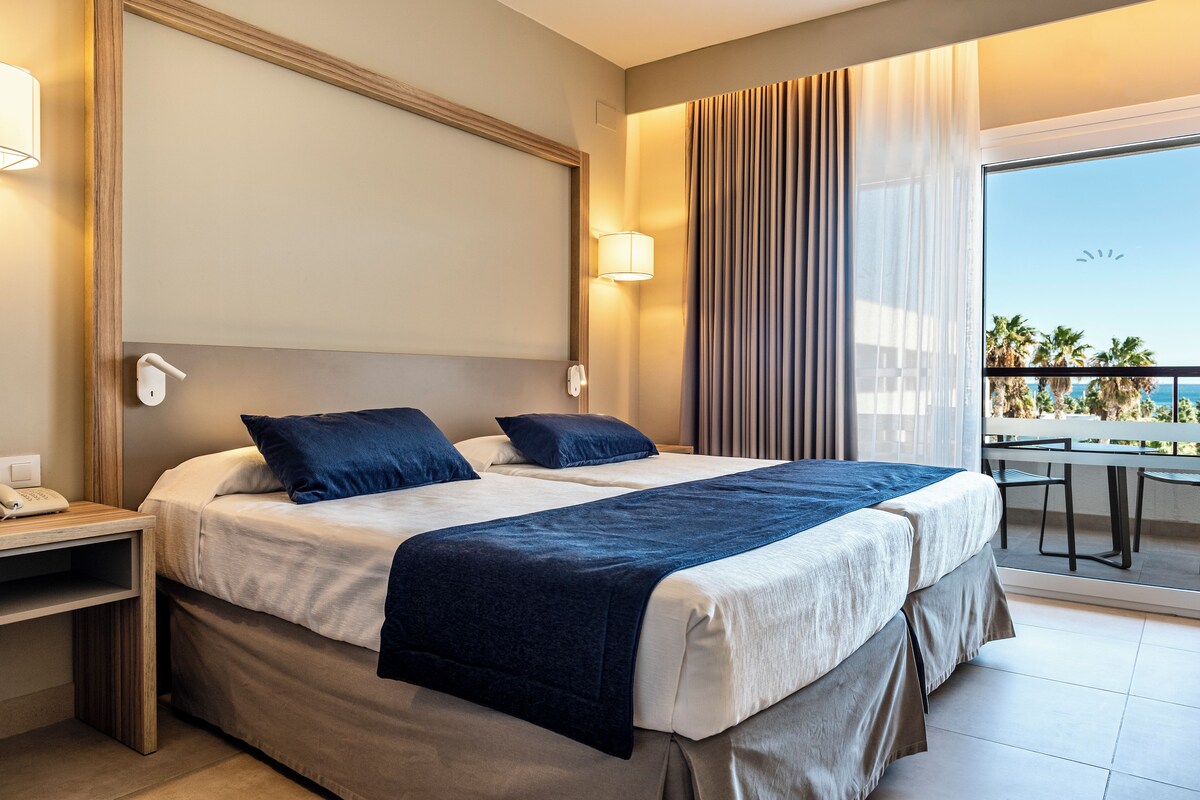 Una habitación de hotel con dos camas, un balcón con vistas, un escritorio con teléfono y una lámpara.