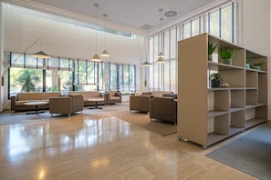 Amplia sala de estar con sofás y una mesa de centro, ventanas grandes con persianas y un estante con plantas.