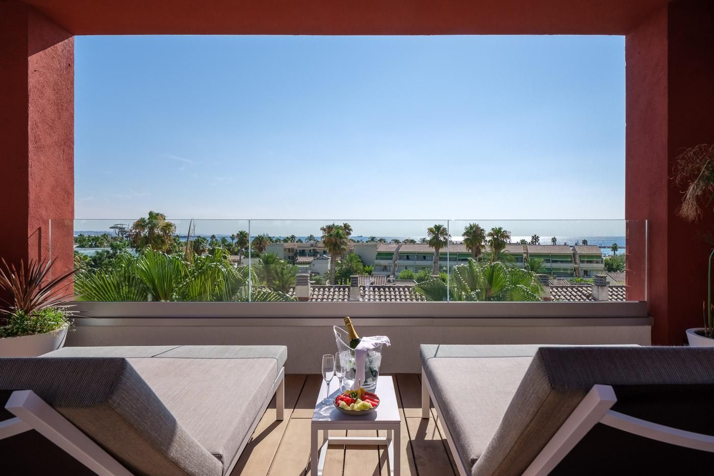 Un balcon avec deux chaises, une table avec une bouteille et des verres, et une vue sur la mer et les bâtiments.