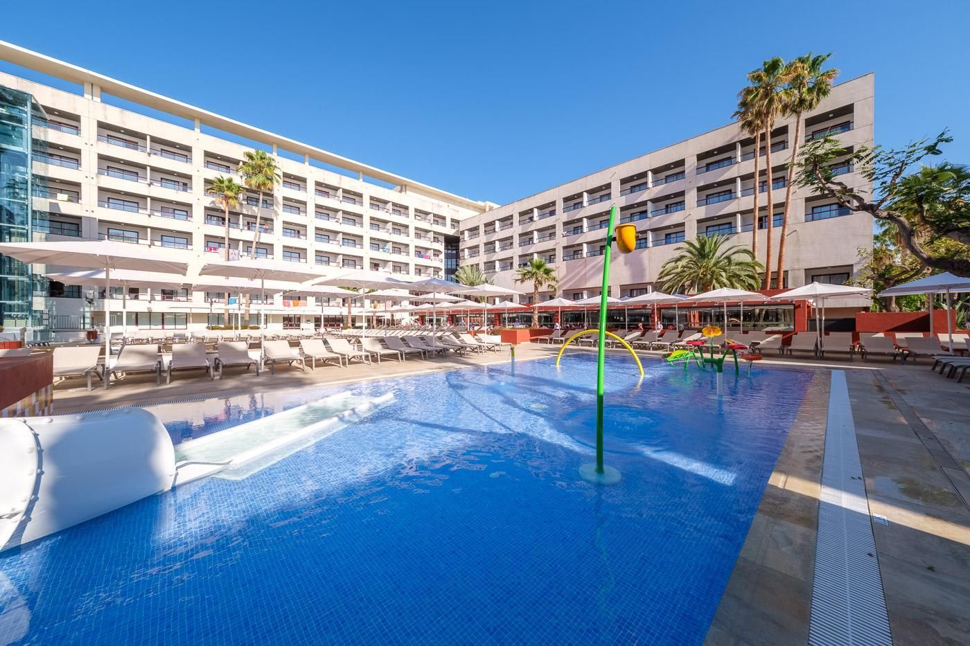A hotel pool area with multiple lounge chairs and umbrellas, surrounded by a multi-story building with balconies.