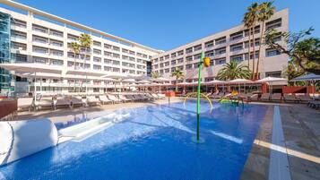 A hotel pool area with multiple lounge chairs and umbrellas, surrounded by a multi-story building with balconies.