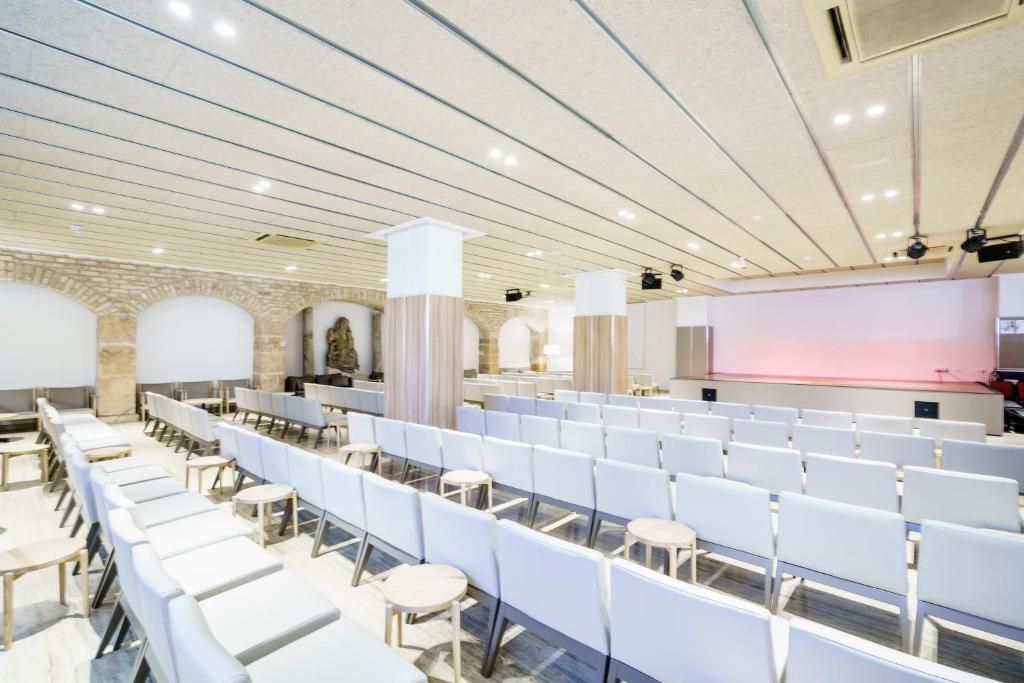 A conference room with rows of white chairs and tables, a stage with a pink backdrop, and a stone wall with arches.