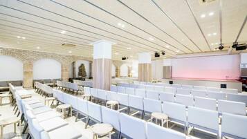 A conference room with rows of white chairs and tables, a stage with a pink backdrop, and a stone wall with arches.
