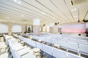 Un salón de conferencias con filas de sillas y mesas blancas, un escenario con un fondo rosa y una pared de piedra con arcos.