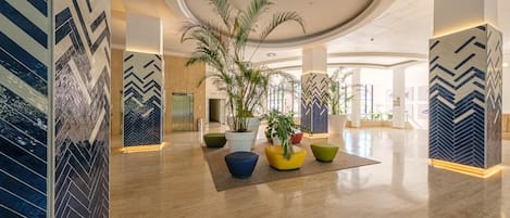 Un'ampia hall dall'arredamento moderno, con un'elegante composizione floreale al centro, un'imponente pianta in vaso e l'ascensore ben visibile.
