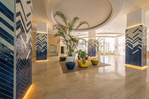 A spacious lobby with a modern design, featuring a central plant arrangement, a large potted tree, and a visible elevator.