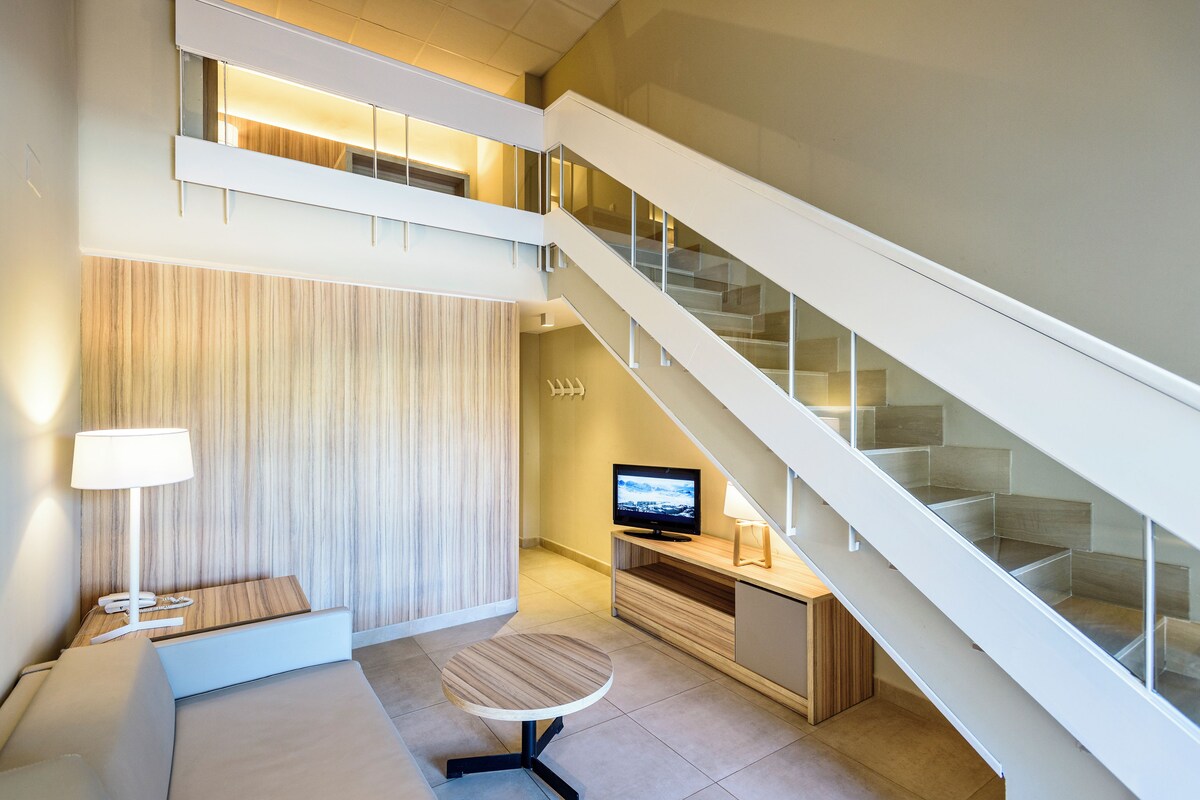 Una habitación de hotel moderna con sofá, una mesita redonda, televisor de pantalla plana y una escalera.