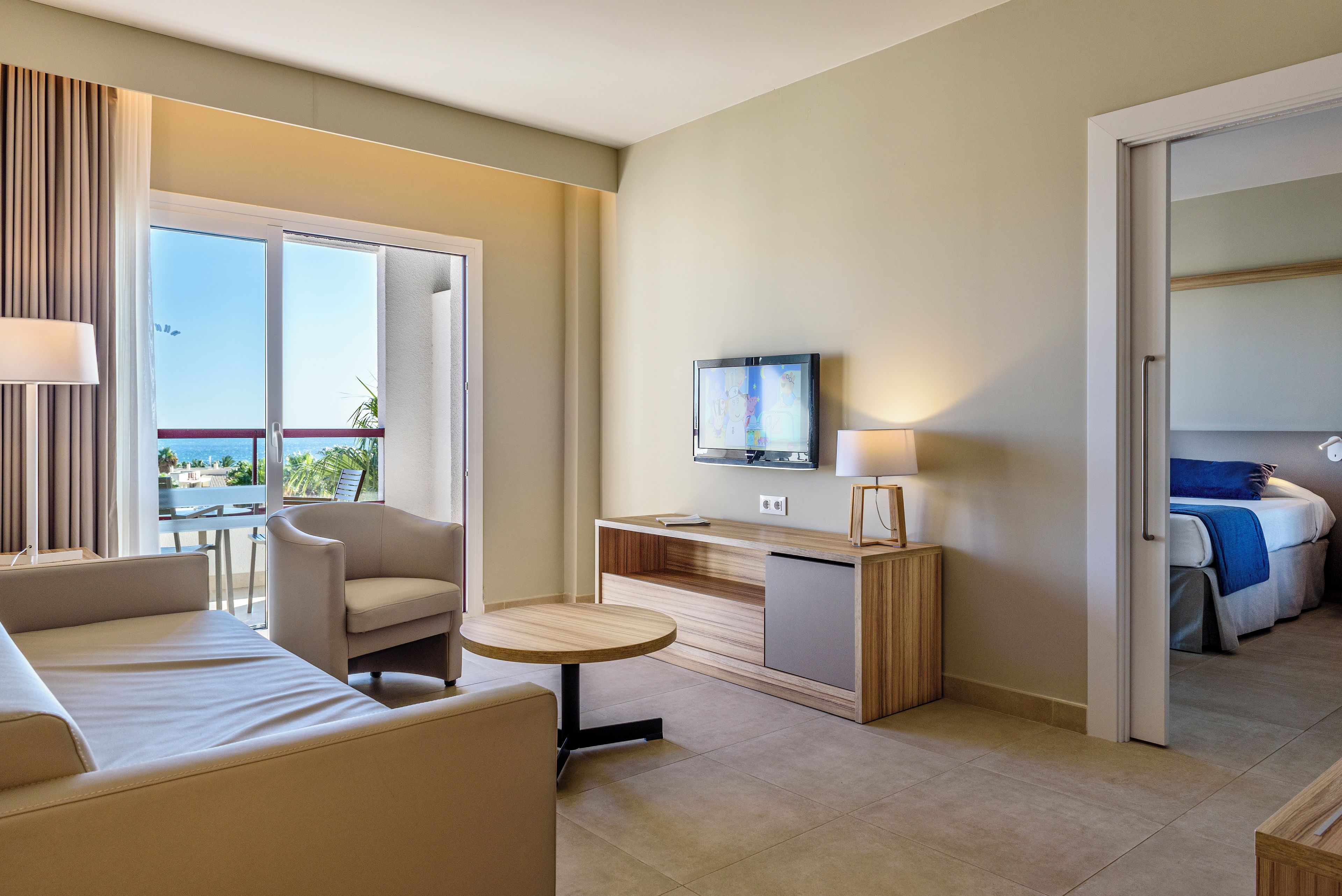 Ein Hotelzimmer mit einer Couch, einem kleinen runden Tisch, einem Fernseher auf einem Holzbalken und einem Bett mit blauer Decke.