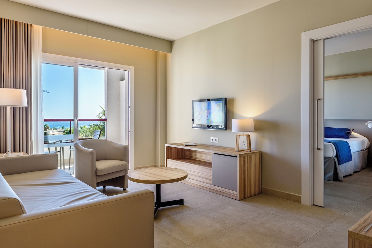 Habitación de hotel moderna con sofá, una mesita redonda, televisor y balcón con vistas.