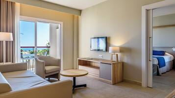 A hotel room with a sofa, a small round table, a TV on a wooden stand, and a bed with a blue blanket.