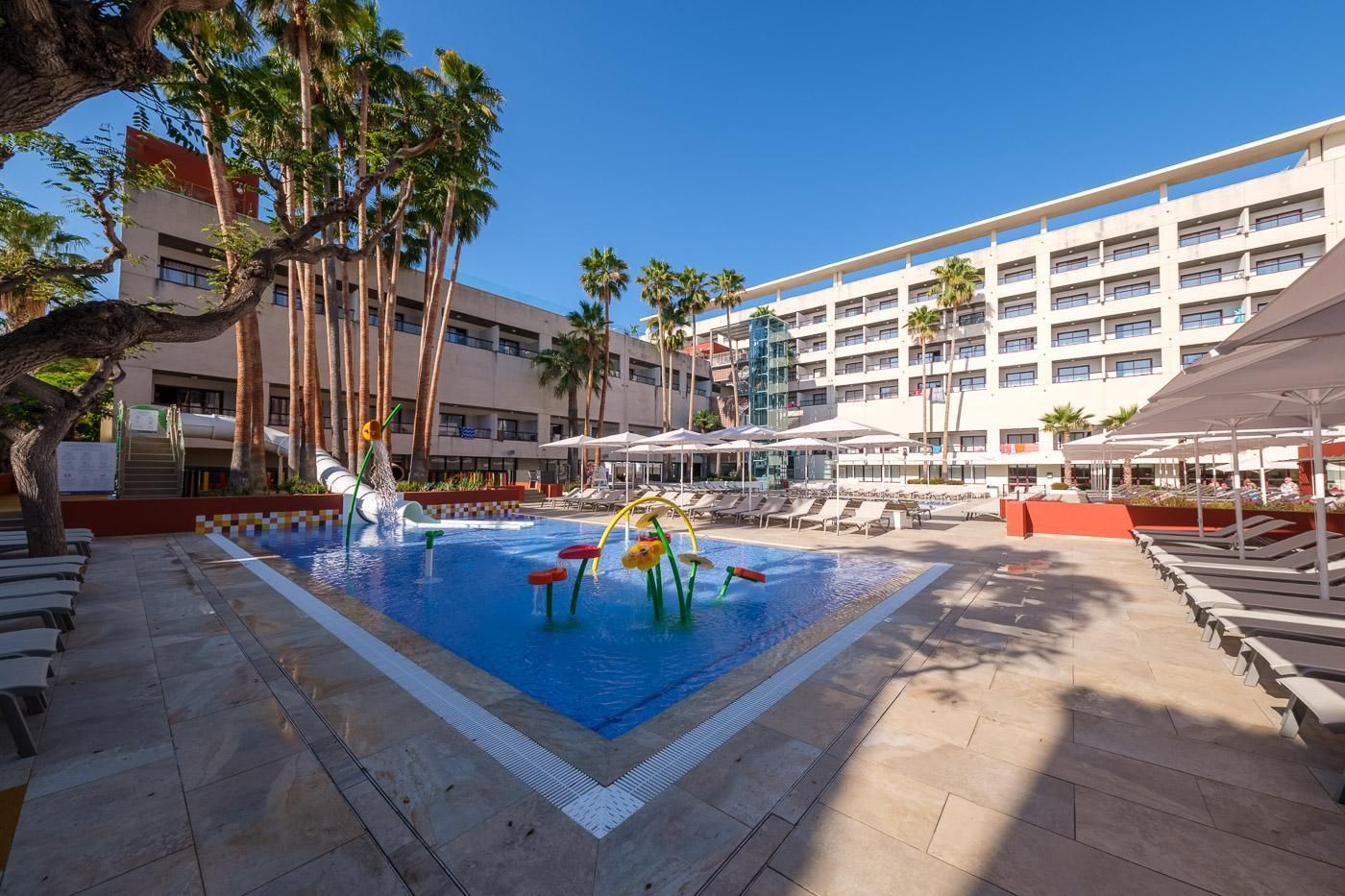 Un espace piscine d’hôtel avec un toboggan aquatique, des chaises longues et des palmiers.