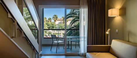 Habitación de hotel con sofá, una mesa, una silla, una escalera y un balcón con vistas a palmeras y una piscina.