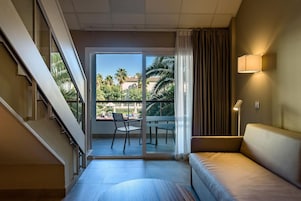 Habitación de hotel con sofá, una mesa, una silla, una escalera y un balcón con vistas a palmeras y una piscina.