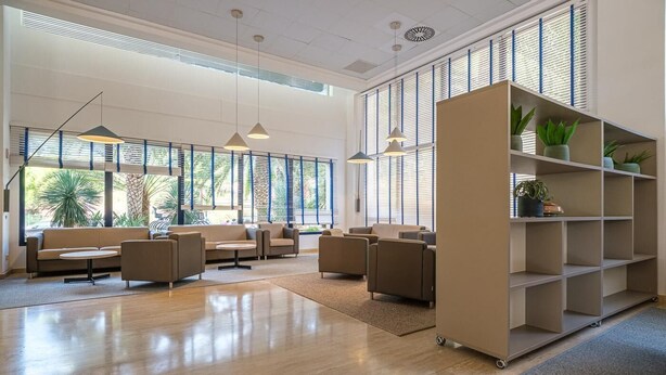 Amplia sala de estar con sofás y una mesa de centro, ventanas grandes con persianas y un estante con plantas.