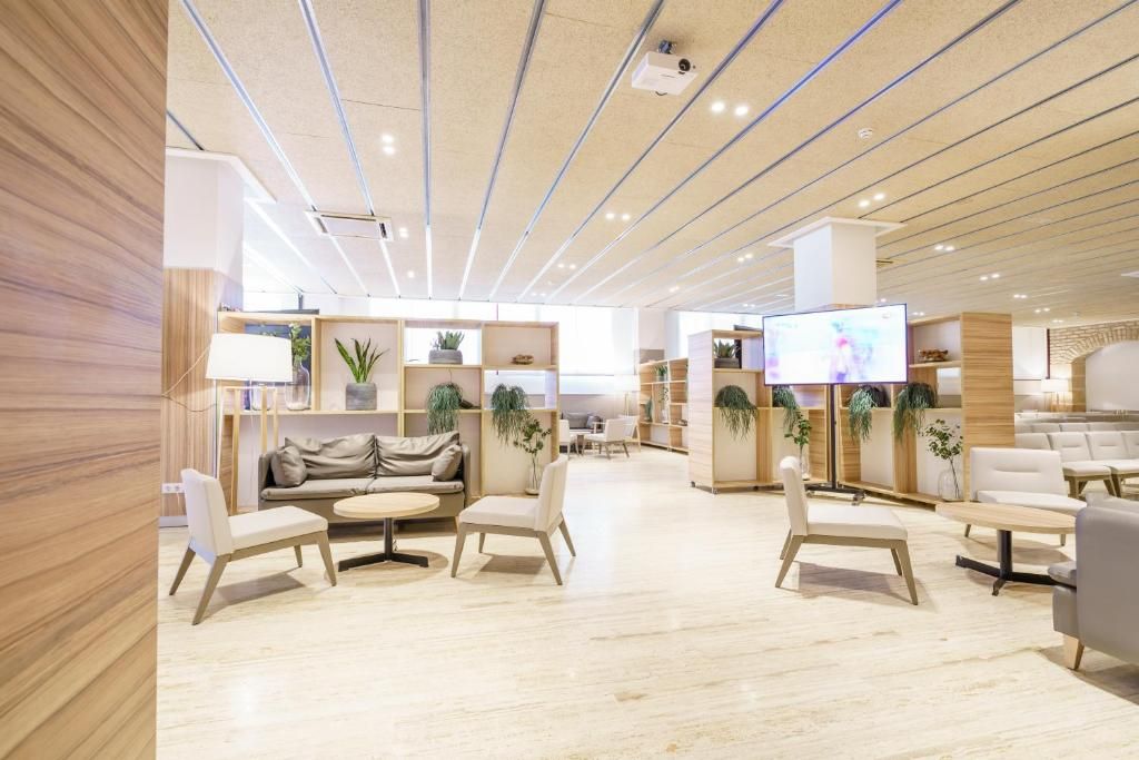 Un hall d’hôtel moderne avec des panneaux de bois, un espace de détente central agrémenté de canapés et de fauteuils, un écran de télévision et des plantes en pot.
