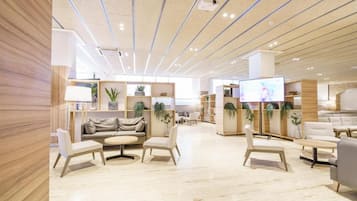 A modern hotel lobby with a central seating area, a television screen, and a ceiling with linear lighting.
